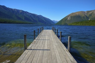 A long wooden jetty juts out into a calm lake, surrounded by mountains under a clear blue sky,
