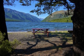A shaded picnic table on the shore of a lake, with mountains and clear sky in the background,