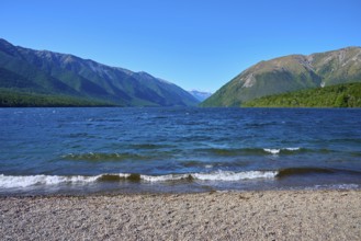 A peaceful lake with gentle waves and a rocky shore, framed by high mountains under a blue sky,