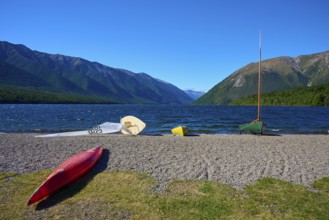 Stored kayaks lying on a beach with a lake and mountains in the background under a clear sky,