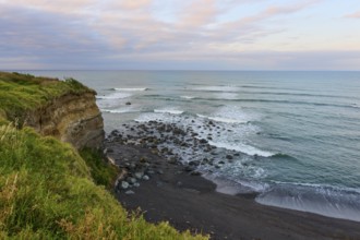 Rocky cliffs by the sea with waves and green vegetation under a cloudy sky, summer, Opunake,