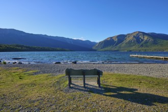 A park bench on the shore of a lake with a view of the surrounding mountains and a boardwalk under