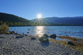 The sun casts dazzling rays over a peaceful lake, framed by rocky shores, summer, Lake Rotoiti,