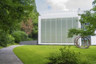 Modern building with a green façade, surrounded by nature and a sculpture in front. A path leads
