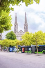 Gothic church with towers behind trees, buses stop on a busy street, Baden Baden, Germany