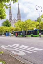An urban street with buses, flanked by trees, gothic church towers visible in the background, Baden