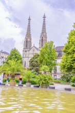 Gothic church with towers in the background, a fountain and palm trees in the foreground, people