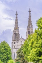 Two tall Gothic church towers rise against a cloudy sky, framed by green trees, Baden Baden,