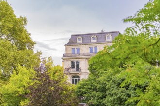 Classic building with numerous windows behind dense trees on a cloudy day, Baden Baden, Germany