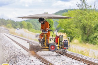 Worker in orange overalls operating a machine on railway tracks, protected by a large umbrella,