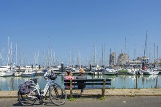 Couple of cyclists resting on bench looking over sailing boats in marina of seaside resort Breskens