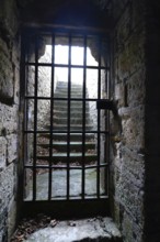 A medieval dungeon in France features thick stone walls and a barred window revealing illuminated