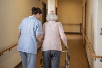 Female nurse helping older woman walk with walker in rehabilitation center hallway. Elder care and