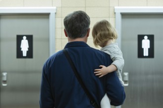 Back view of father with young daughter in front of men's and women's restroom doors. Concept of