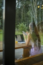 Rear view at a woman working in the evening outdoors while sitting on the terrace of a log cabin