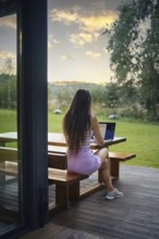 Rear view of an unrecognizable woman sitting on terrace in the evening and typing on laptop
