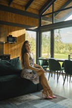 Happy woman in a floral dress sits on a couch in a sunlit room with large windows and contemporary