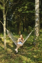 A relaxed woman reads message on smartphone while lounging in a hammock amidst lush green woods