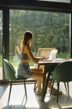 Woman using a laptop at a modern desk near a large window with greenery outside