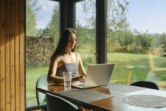 A focused woman working on a laptop in a calm cabin environment with a view of nature