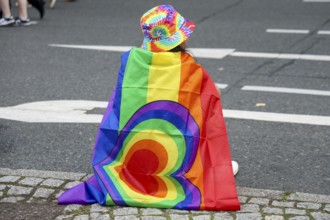A participant with a rainbow flag and colourful hat sits on the side of the road at the 47th
