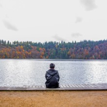 A solitary figure sits on a wooden dock, looking out over a tranquil lake. Autumn trees reflect