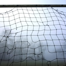A weathered football goal net looms in the foreground surrounded by dense fog, with a blurred view