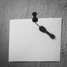 A blank piece of note paper is pinned to a textured wall, casting a compelling shadow that