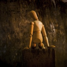 A wooden mannequin is positioned on a rough wooden surface, looking contemplative against a