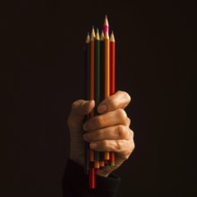 A hand grasps a variety of colored pencils, showcasing the range of colors. The scene is