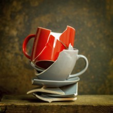 Stack of broken red mug and gray plate pieces on rustic wooden surface, showcasing fractured