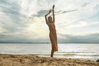 A woman in a floral sundress stands barefoot on the sand, joyfully lifting her arms. The sun sets