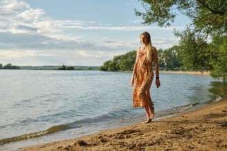 A woman in a colourful sundress strolls barefoot along the sandy shore of a tranquil lake. Lush