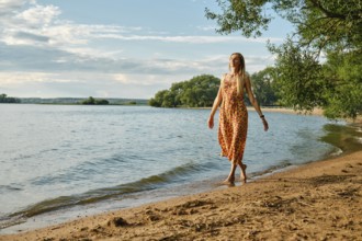 A woman in a vibrant sundress strolls barefoot along the sandy beach beside a calm lake. Sunshine
