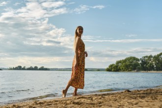A woman is strolling barefoot along a sandy riverbank, wearing a colourful floral sundress. The