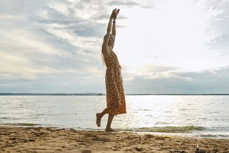 A woman in a colourful sundress walks along a serene lake, raising her arms in joy. The cloudy sky