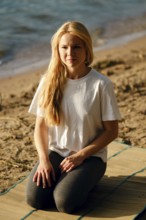 A young woman sits cross-legged on a mat at the beach, gazing thoughtfully towards the water. The
