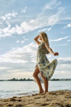 A woman stands gracefully on the sandy shore, her floral dress flowing in the wind. She gazes