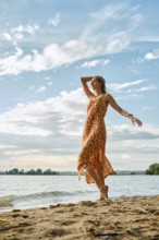 A woman stands barefoot on sandy shores, wearing a bright dress with a bold floral pattern. She