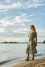 A woman wearing a floral dress stands barefoot on the sandy shore of a lake, gazing at the water.