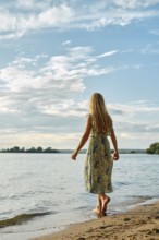 Rear view of barefoot woman standing on a sandy beach by a tranquil lake. The clear blue sky
