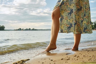 Unrecognizable woman stands at the edge of a lake, her bare foot touching the gentle waves at