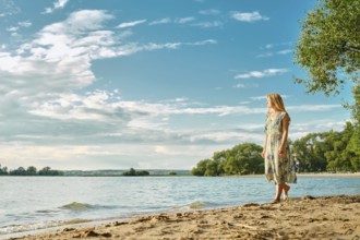 A woman in a floral dress strolls barefoot along the sandy shore of a peaceful lake, surrounded by