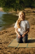 A woman sits on a mat by the riverside, engaged in a moment of mindfulness. She has long blonde