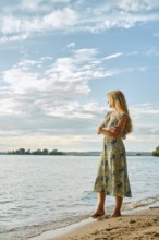 Thoughtful woman with long blonde hair stands on the sandy shore, gazing thoughtfully at the water