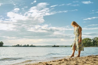 A woman wades along the sandy shore, wearing a colorful floral dress. The sun shines brightly, and