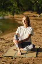 A woman sits cross-legged on a mat by a riverbank, focusing on mindfulness as the sun sets. She