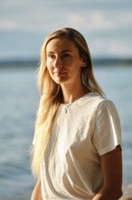 A woman with long blonde hair is standing near the water's edge at sunset. She gazes thoughtfully
