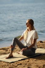 A woman sits on yoga mat by the lakeshore and listens a surf noise. The calm water reflects the