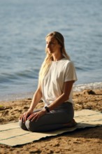 A woman sits calmly on a yoga mat by the water, practicing mindfulness as the sun sets. The serene